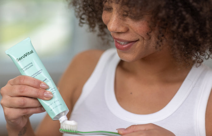 Woman applying toothpaste to a toothbrush with a tube labeled SeroVital in the foreground.