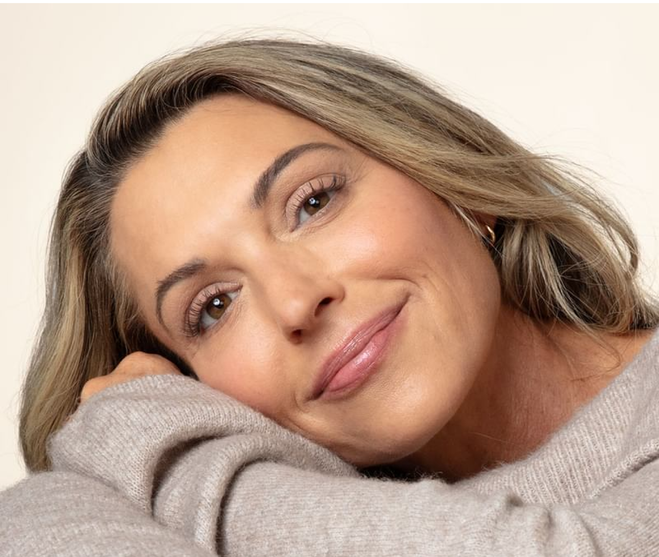 This is a close-up portrait of a woman with shoulder-length, light brown hair, smiling softly and resting her chin on her hand.