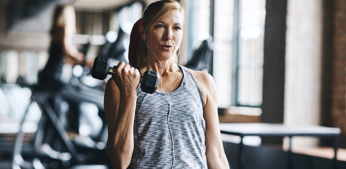 A midlife woman wearing earbuds and a gray workout top performing a dumbbell biceps curl at the gym.