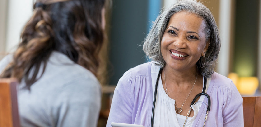 A cheerful senior woman wears a stethoscope as she sits with an unrecognizable woman during an interview. She holds a digital tablet.