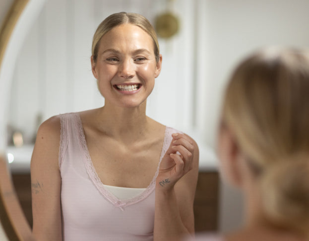 Woman smiling at herself in the mirror wearing a pink tank top.
