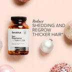 A glass bottle of hair supplement Hair Regeneres ADVANCED on a pink background with holiday ornaments and text that says Reduce shedding and regrow thicker hair