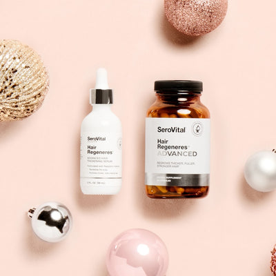 A bottle of Hair Regeneres advanced thickening serum and a glass bottle of Hair Regeneres ADVANCED hair regrowth supplement on a pink background with holiday ornaments