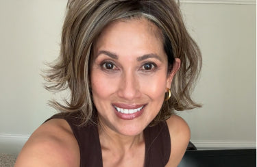 A woman with shoulder-length, wavy blonde and brown hair, wearing a sleeveless brown top, smiles at the camera.