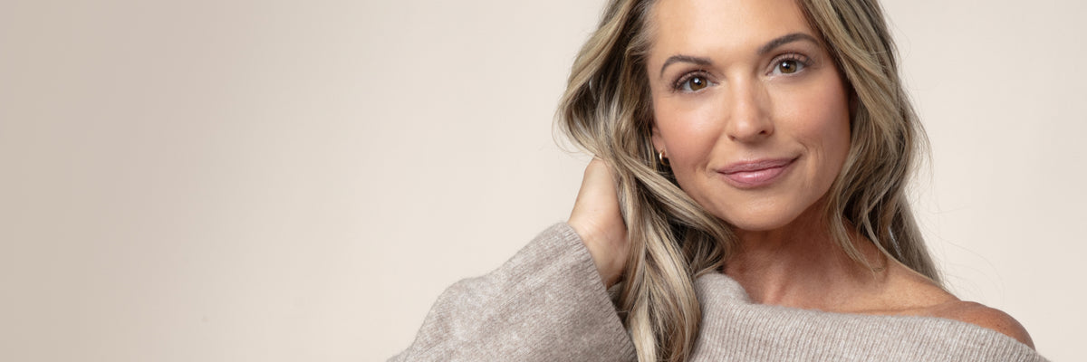 A woman with blonde hair wearing a beige off-the-shoulder knit sweater, posing against a solid light beige background.