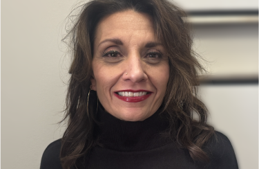 A woman with shoulder-length brown hair, wearing a black turtleneck sweater, smiles at the camera. She has red lipstick on and is wearing gold hoop earrings.
