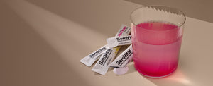 A glass of pink liquid next to several SeroVital Rapid Dissolve Powder stick packs, with some powder spilled out.