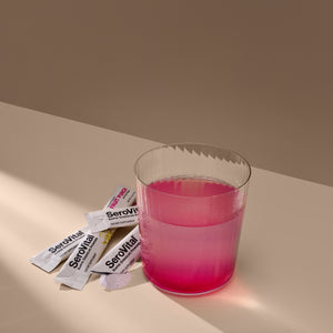 A glass of pink liquid next to several SeroVital Rapid Dissolve Powder stick packs, with some powder spilled out.