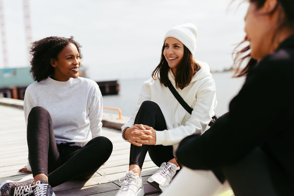 Three happy women outside in activewear sitting next to each other at a pier