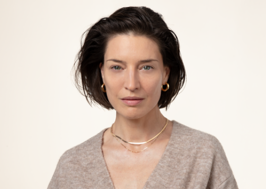 This is a portrait of a woman with short, dark brown hair and blue eyes, wearing a beige sweater, gold hoop earrings, and a delicate gold necklace.