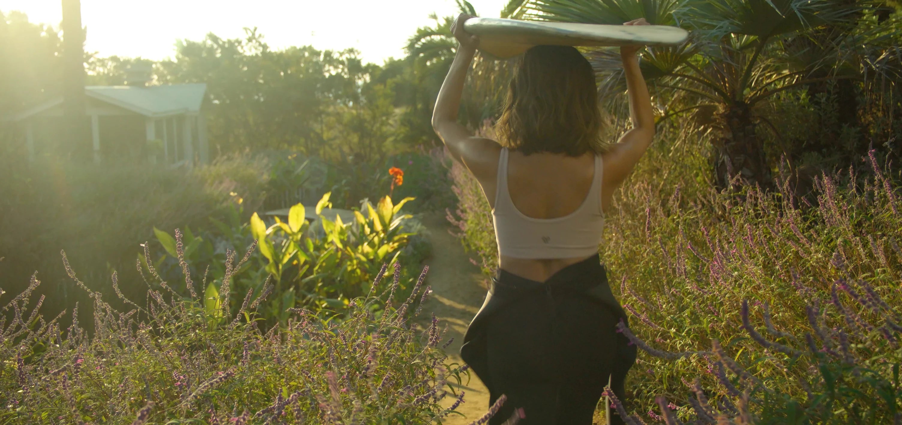 A woman with shoulder-length brown hair, wearing a light-colored tank top and black pants, walks along a path in a lush garden. She carries a surfboard above her head with both hands.
