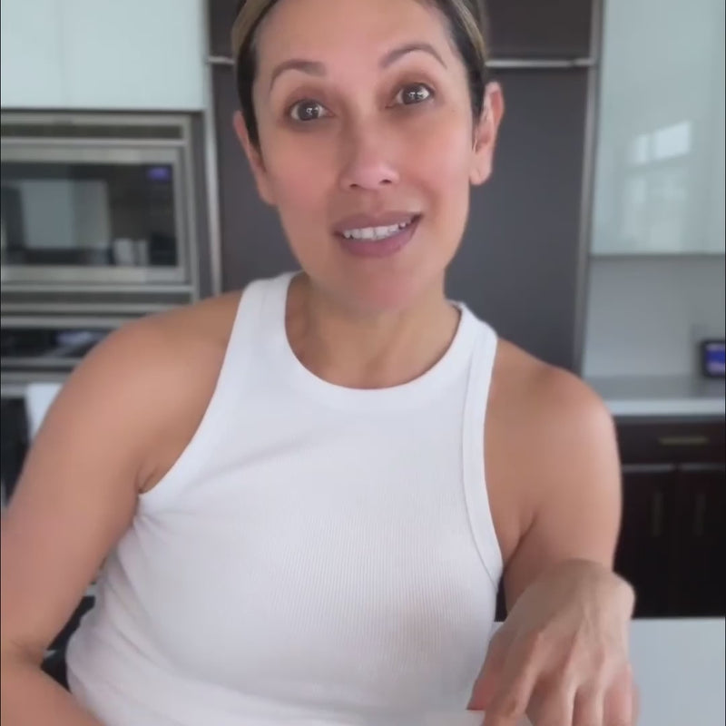 A gorgeous, fresh-faced woman with her hair in a ponytail, wearing a white tank top, holding a box of anti-aging powder Glow Sticks, then drinking a glass of it
