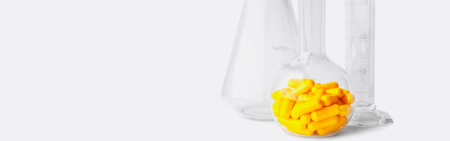 Close-up photo of a clear glass flask filled with bright yellow capsules, with a blurry conical flask and a graduated cylinder in the background, all on a white surface.