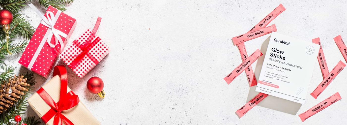 Serovital Glow Sticks Beauty Illumination box and individual packets on a white, holiday-themed background with red and white wrapped Christmas gifts and evergreen decor. A perfect holiday gift idea.