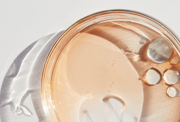 A close-up image of a clear glass dish containing a light peach-colored liquid with several clear droplets floating on the surface.