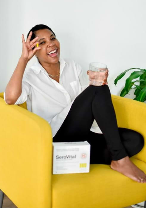 A black woman with very short, dark hair sitting on a yellow couch smiling and holding out a single pill of the anti-aging breakthrough SeroVital